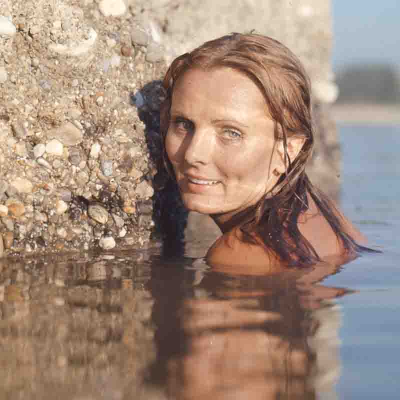 Eine Nahaufnahme der deutschen Schauspielerin Ruth Maria Kubitschek, fotografiert von Ingeborg Bock-Schroeder. Sie befindet sich schultertief im Wasser und schaut lächelnd mit nassen, braunen Haaren in die Kamera. Sie lehnt sich an einen von Algen und Kieselsteinen bewachsenen Felsen oder Wellenbrecher, dessen Spiegelung im ruhigen Wasser zu sehen ist.